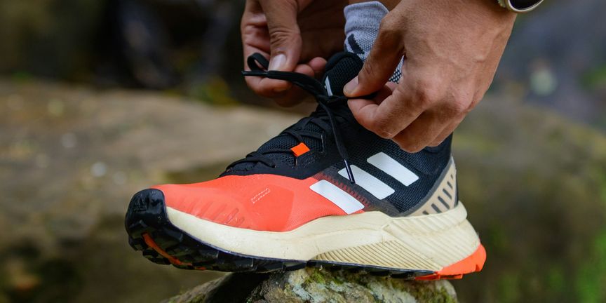 Handen strikken de veters van een oranje-zwarte trailschoen op een mossige steen, met een gps-horloge om de pols