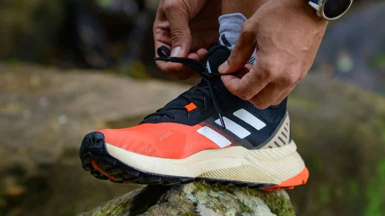 Handen strikken de veters van een oranje-zwarte trailschoen op een mossige steen, met een gps-horloge om de pols