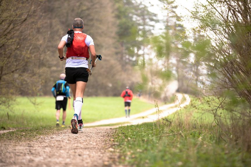 Trailrunner van achteren met stokken op een grindpad door groene velden, twee lopers verderop