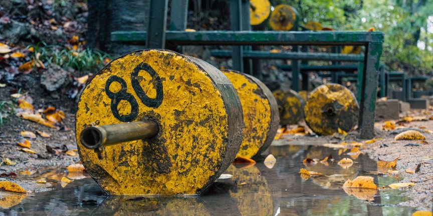 Verweerde gele halter met '80' erop, liggend in de modder bij een openluchtgym in het bos