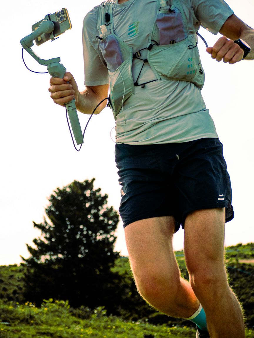 Hardloper met hydratievest, zachte flesjes en een gps-horloge, die een telefoon op een gimbal vasthoudt tijdens het lopen.