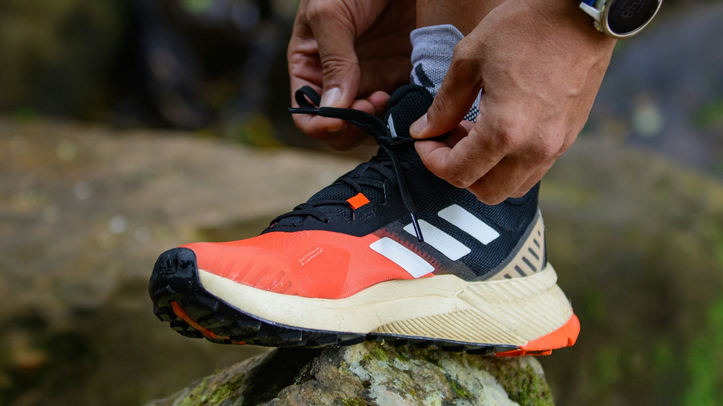 Handen strikken de veters van een oranje-zwarte trailschoen op een mossige steen, met een gps-horloge om de pols