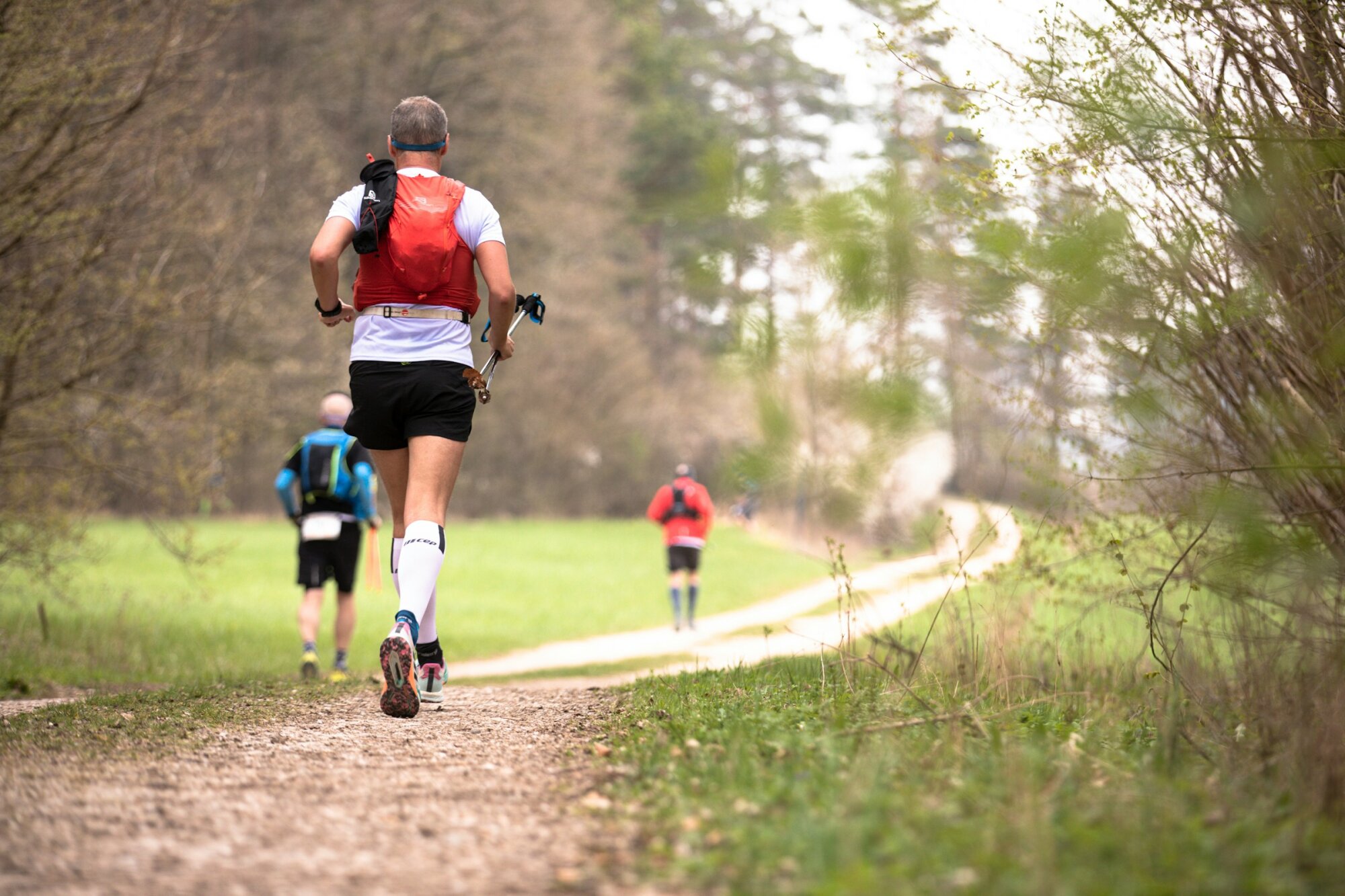 Trailrunner van achteren met stokken op een grindpad door groene velden, twee lopers verderop